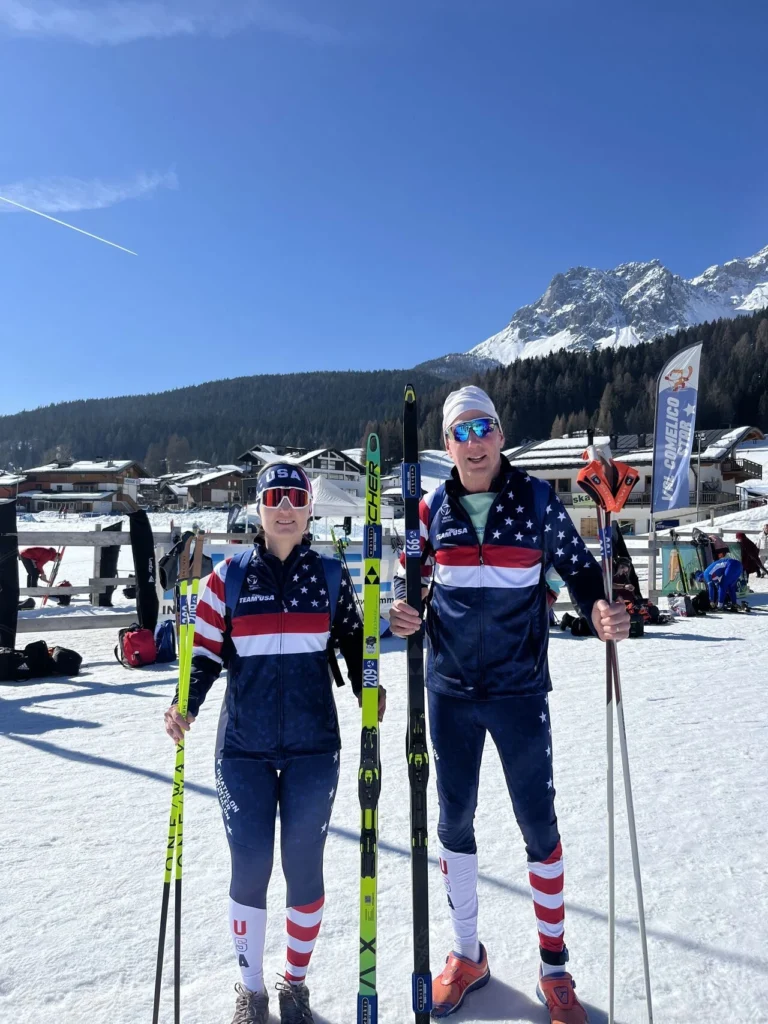 A man and a woman holding skiing gear with snowy mountains in the background, wearing USA outfits